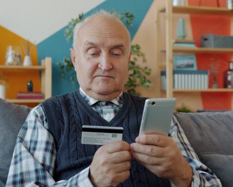 Elderly man holding credit card and smartphone for online shopping.