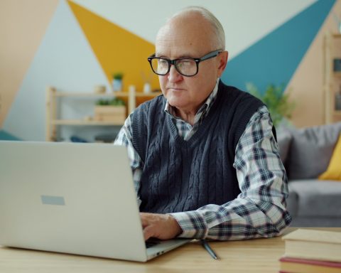 Elderly man wearing glasses using a laptop at home.