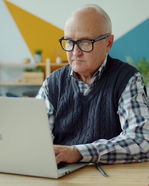 Elderly man wearing glasses using a laptop at home.
