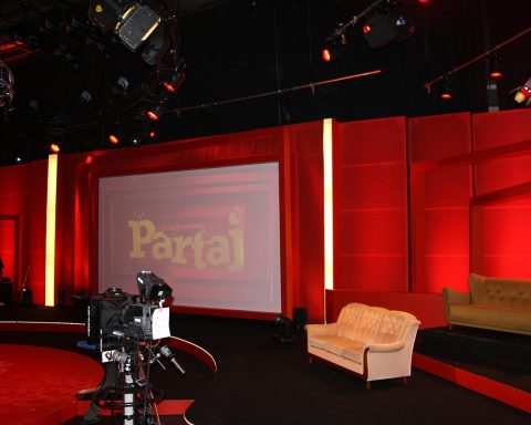 Een oudere man met een doorleefde uitstraling zit in een televisiestudio, klaar voor een interview