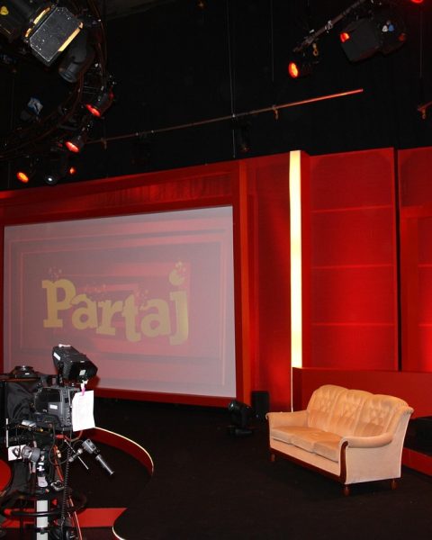 Een oudere man met een doorleefde uitstraling zit in een televisiestudio, klaar voor een interview
